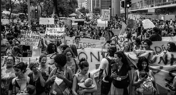 Black and white image of a demonstration with many women in the foreground carrying babies in slings and others holding banners and placards.